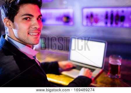 Portrait of smiling man using laptop with glass of beer on table at bar counter in bar