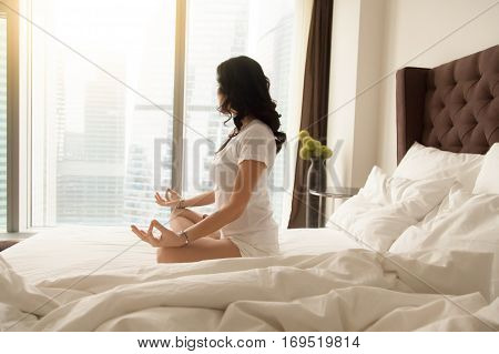 Young sporty woman practicing yoga at home bedroom after waking up, sitting on unmade bed in Padmasana exercise, Half Lotus pose on meditation session, working out wearing white casual clothes, indoor