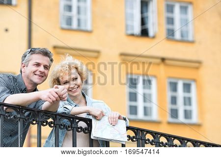 Happy middle-aged couple with map leaning on railing against building