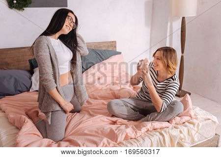 Look at the camera. Attentive committed beautiful lady being a personal photographer while the other girl posing for her in a comfy home outfit and enjoying the process