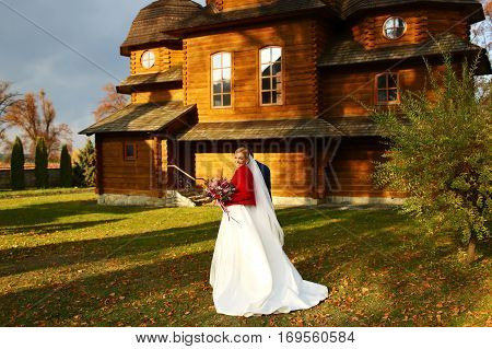 Bride Smiles Standing With A Weding Bouquet On The Backyard Of An Old Wooden Church