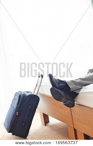 Low section of businessman lying on bed beside luggage in hotel room