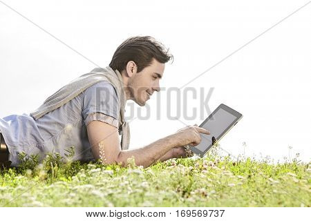 Side view of young man using digital tablet while lying on grass against clear sky