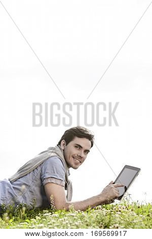 Side view portrait of young man using digital tablet while lying on grass against clear sky