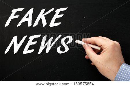 Male hand writing the words Fake News in white text on a blackboard as a reminder to be aware of hoaxes and disinformation for propaganda uses