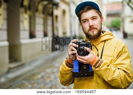 Young hipster with a beard holding oldshool film camera, thinking and ready to take a photo, on the street, wearing in bright raincoat and hat, portrait, day outdoors, closeup, sly glance
