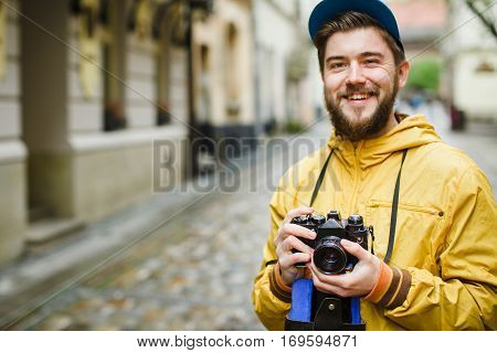 Young hipster with a beard holding oldshool film camera, laughing and ready to take a photo, on the street, wearing in bright raincoat and hat, portrait, day outdoors, closeup, soft-focus