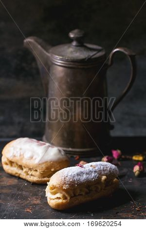 Two eclairs with white chocolate and vintage teapot over dark background.