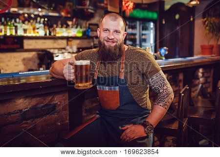 Bearded barman with tattoos wearing an apron sitting near the bar and holding a glass of beer