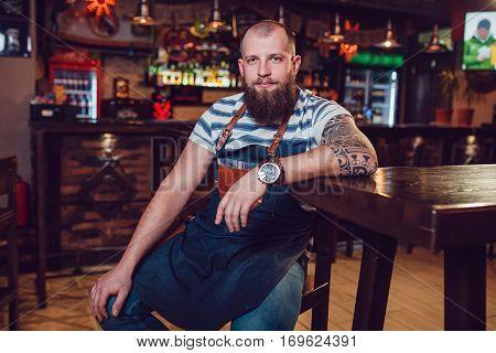 Bearded barman with tattoos and wathes wearing an apron sitting in bar.