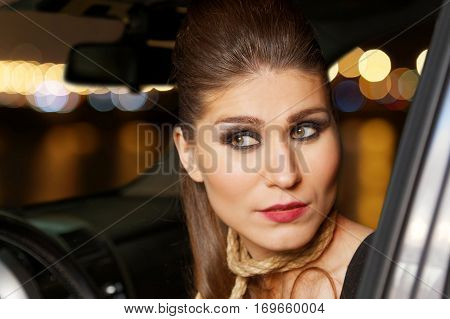 Attractive sexy young woman with an evening make-up with a rope around his neck instead of necklaces sitting in the car. Lights of night city on blurred background.