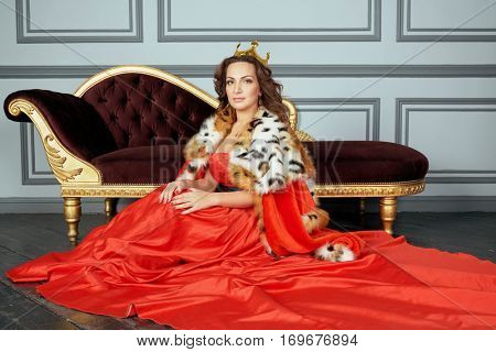Woman in red dress, cloak and with crown on head sits on floor near couch in room.