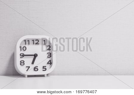 Closeup white clock for decorate show a quarter to seven p.m. or 6:45 a.m. on white wood desk and cream wallpaper textured background in black and white tone with copy space