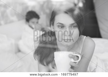 Thougtful woman having coffee at home with man lying in background