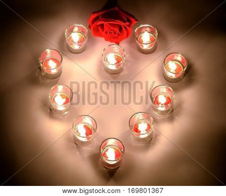 A small lighting lamps with red color aromatic paraffin in a small glasses arrange in heart form with a red rose symbol of love and valentine's day on a top of the heart put on a white floor in the night