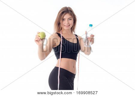 Beautiful slim girl in a black top and tights is turned sideways and stretched out both hands forward shows a fruit and a bottle of water and smiling.