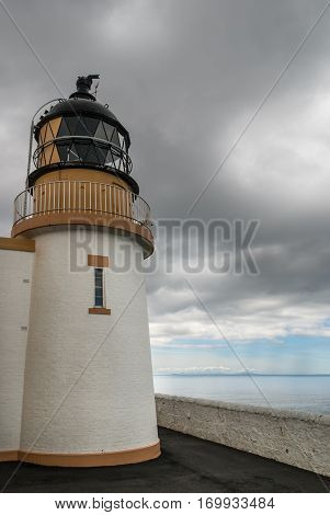 Assynt Peninsula Scotland - June 7 2012: The white tower of lighthouse complex at Stoer Head has a golden rim with the black light technology part on top. Gray cloudscape and light blue ocean water.