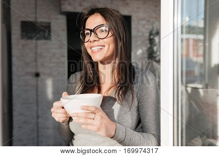 Photo of young beautiful lady wearing eyeglasses drinking coffee indoors. Look aside.