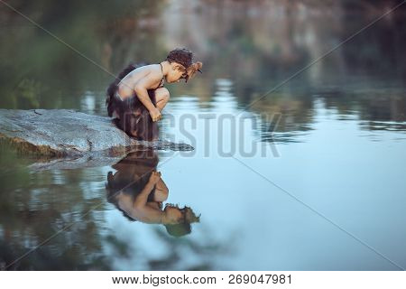 Caveman Boy Sitting On The Rock And Looking At Him Self In The Water Reflection In Lake. Evolution S