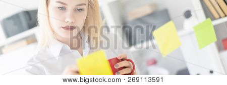 A Young Girl Stands Near A Transparent Board With Stickers. Shes Holding A Cup And A Marker.