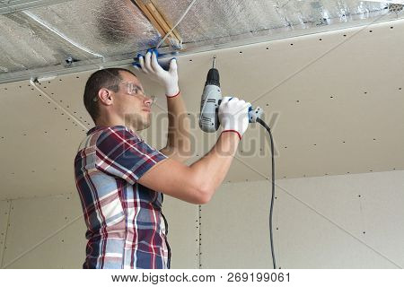 Young Man In Goggles Fixing Drywall Suspended Ceiling To Metal Frame Using Electrical Screwdriver On