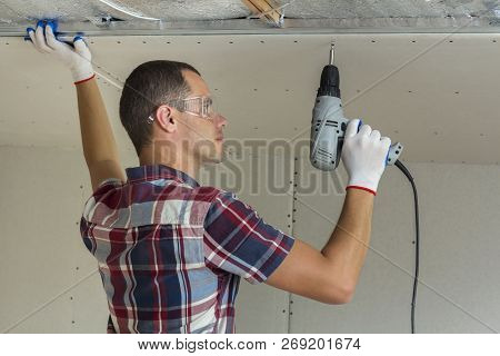 Young Man In Goggles Fixing Drywall Suspended Ceiling To Metal Frame Using Electrical Screwdriver On