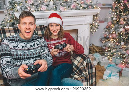 Two People Sitting And Playing Games. Thay Hold Gamepads In Hands. Young Man And Woman Are Excited. 