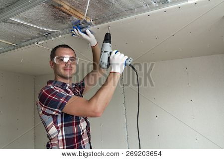 Young Man In Goggles Fixing Drywall Suspended Ceiling To Metal Frame Using Electrical Screwdriver On