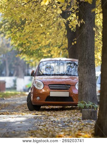 Front View Of Small Orange Mini Car Parked In Quiet Yard On Sunny Autumn Day On Blurred Buildings An