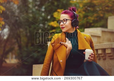 Cute Young Girl Listening To Music On Headphones In Autumn Park. Woman Is Holding Paper Cup With Hot