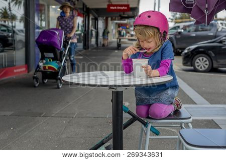 Young Girl In Pink Outfit And Pink Helmet Riding A Kick-scooter Sitting At The Outside Cafe Table Wi