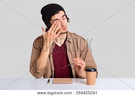 Tired Hipster Rubbs Eyes As Wants To Sleep, Sits At Workplace With Notepad, Pen And Coffee, Wears Ca
