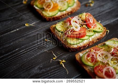 Row of three healthy toasts with onion, cucomber, cherry tomatoes, crumbled feta cheese and radish sprouts on a black plate. Table top view on black wooden background