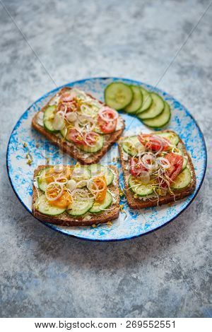 Plate with healthy toasts with cucomber, cherry tomatoes, crumbled feta cheese and radish sprouts on a black plate. Table top view. Gray stone background.
