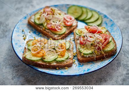 Plate with healthy toasts with cucomber, cherry tomatoes, crumbled feta cheese and radish sprouts on a black plate. Table top view. Gray stone background.
