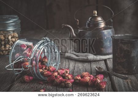 Vintage Teapot, Cup Of Healthy Herbal Tea, And Glass Jars Of Dry Rose Buds And Daisy Medicinal Herbs