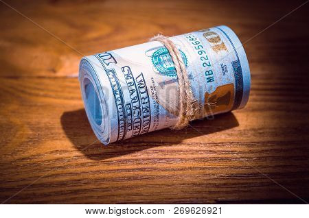 A Roll Of Dollars On A Wooden Textured Table In The Dark Illuminated By A Ray Of Light.