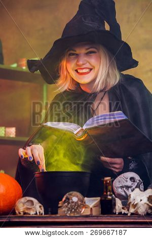 Photo of smiling witch blonde in black hat with book potting potion in pot