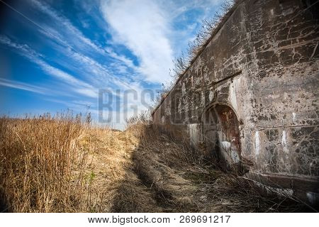 Old Destroyed Wall With Deepening In The Field Of Dried Grass And Blue Sky With Clouds.