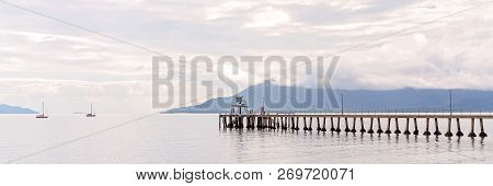 Wooden Pier Jutting Out Into The Ocean On A Calm Day With Mountain Background And Cloudy Sky