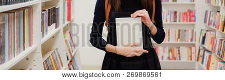 Female Hands Holding A Book In Shop On Background Of Bookshelves. Girl Choosing What To Read In Book