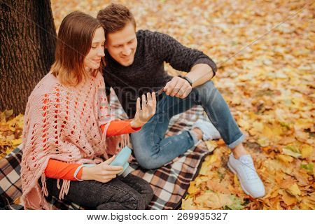Chherful Young Man And Woman Sit On Blanket And Look At Phoen They Hold In Hands. Girl Has Blue Came