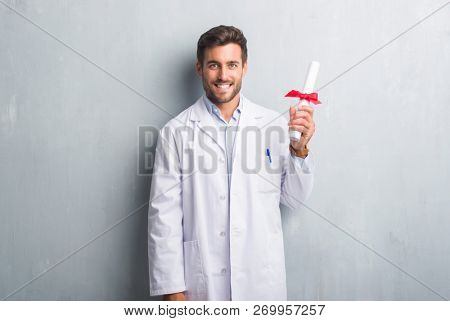 Handsome young doctor man over grey grunge wall holding diploma with a happy face standing and smiling with a confident smile showing teeth