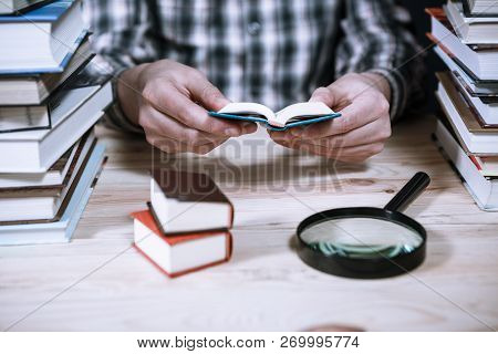 A Man Reading A Miniature Book Among The Big Books In The Library At The Wooden Table.