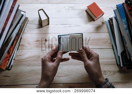 A Man Reading A Miniature Book Among The Big Books In The Library At The Wooden Table.