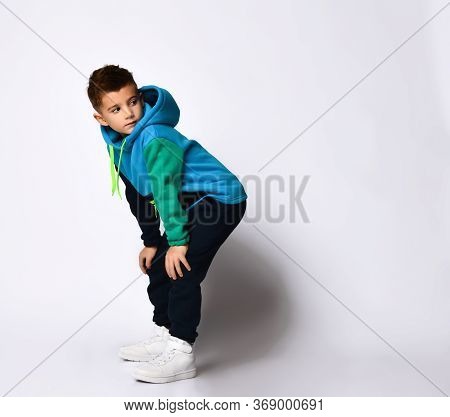 Stylish Little Boy In A Warm Tracksuit Rests His Hands On His Knees And Looks Away.