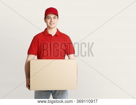 Work Of Modern Postman. Smiling Young Guy In Uniform Holding Big Cardboard Box Isolated On Light Bac