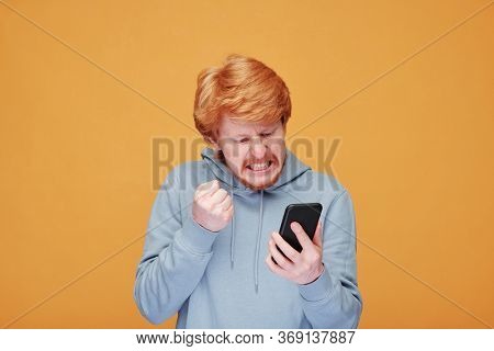 Extremely angry young bearded man in casualwear looking at screen of smartphone while having argument with someone in video chat