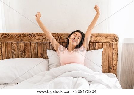 Joyful Asian Woman Waking Up Stretching Hands Enjoying Weekend Morning At Home Sitting In Bed Indoor