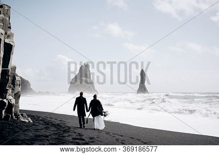 Destination Iceland Wedding. A Wedding Couple Walks Along The Sandy Black Beach Of Vik, Near The Bas